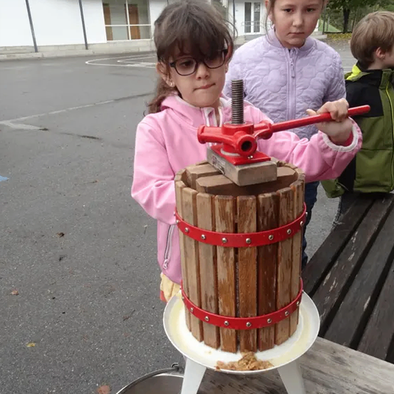 Ein junges Mädchen in einer rosa Jacke bedient eine Holzpresse im Freien. Sie hält einen roten Griff und steht neben einem Fass und einer Bank. Hinter ihr beobachten zwei Kinder.