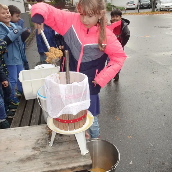 Ein Mädchen benutzt eine manuelle Presse, um Saft aus Äpfeln zu extrahieren. Andere Kinder beobachten im Hintergrund.