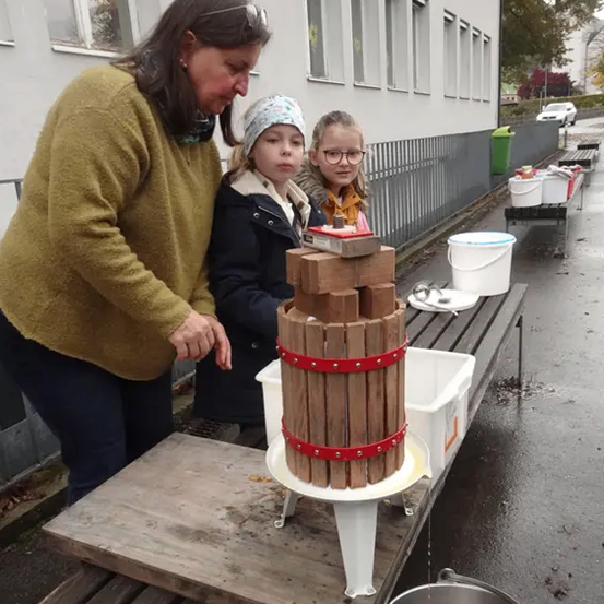 Eine Frau und zwei Mädchen pressen Äpfel in einer hölzernen Presse vor einem Gebäude.
