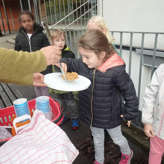 Kinder versammeln sich um einen Tisch, wobei ein Mädchen einen Teller mit Essen hält und ein anderes Mädchen Essen von einer Person erhält. Dahinter befinden sich eine Treppe und ein Gebäude.