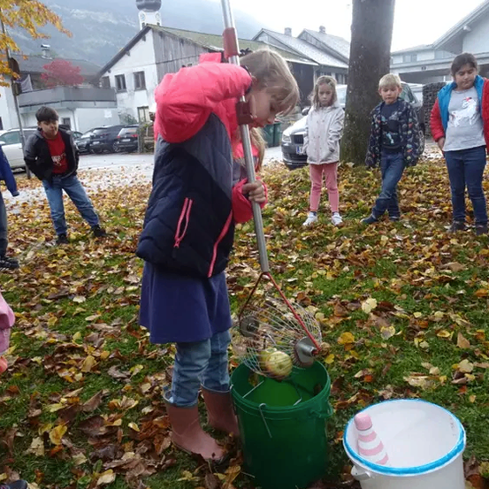 Ein Mädchen in einer rosa Jacke sammelt Äpfel mit einem Rechen in einem grünen Eimer, während mehrere Kinder hinter ihr stehen. Die Szene spielt sich auf einem Außengelände mit verstreuten Blättern und einem bewölkten Himmel ab.