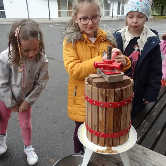 Drei junge Mädchen sind draußen und benutzen eine manuelle Apfelpresse. Ein Mädchen drückt, während zwei andere zuschauen, mit einem Gebäude im Hintergrund.