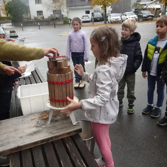 Ein Mädchen drückt eine Holzpresse, während andere zusehen, mit Autos und Gebäuden im Hintergrund.