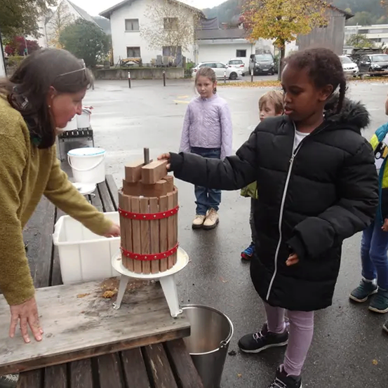 Eine Frau und ein Kind benutzen eine hölzerne Presse im Freien. Andere Kinder und Erwachsene sind im Hintergrund.
