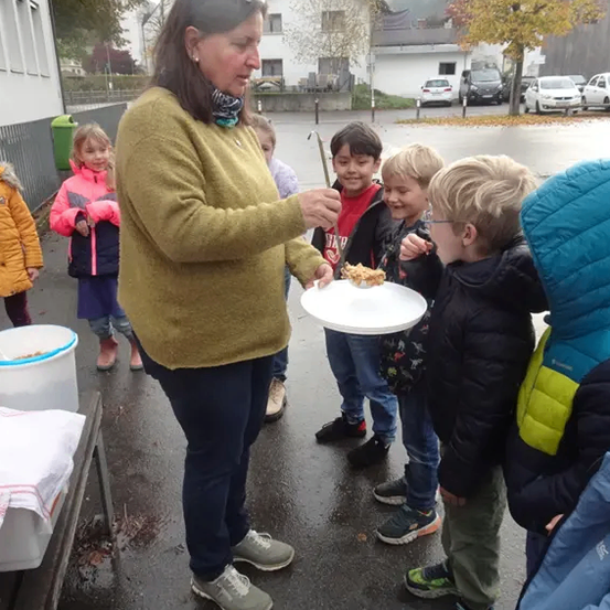 Eine Frau in einem gelben Pullover füttert Kinder mit Essen auf einem Teller. Die Kinder sind in Jacken gekleidet. Auf dem Tisch stehen zwei weiße Eimer.