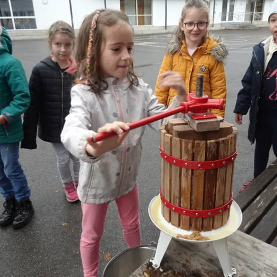 Ein kleines Mädchen benutzt eine rote Holzpresse, während drei andere Kinder zusehen. Sie sind draußen, möglicherweise auf dem Schulhof. Hinter ihnen steht ein Gebäude mit vielen Fenstern.