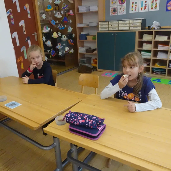Zwei Mädchen sitzen in einem Klassenzimmer und essen. Eines hat ein Stück Brot in der Hand und das andere isst aus einem kleinen Becher.