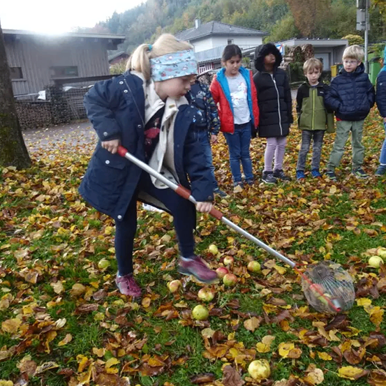 Ein Mädchen mit Stirnband benutzt einen Rechen, um Äpfel auf dem Gras mit gefallenen Blättern aufzuheben. Kinder stehen hinter ihr und beobachten. Im Hintergrund befinden sich Gebäude und Bäume.