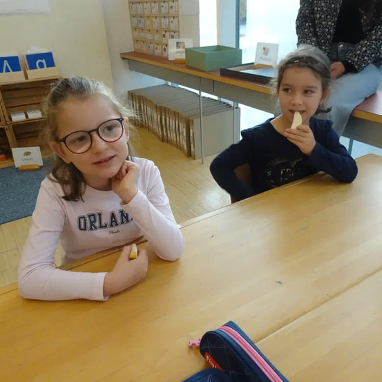 Zwei junge Mädchen in einem Klassenzimmer, eine mit Brille, beide essen ein Stück Essen. Ein Holztisch, ein Stuhl und ein Federmäppchen sind im Vordergrund.