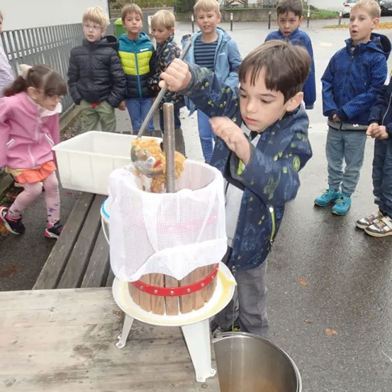 Ein Junge presst Äpfel in einer Presse im Freien, umgeben von anderen Kindern. Er drückt die Früchte mit einem Stab zusammen und Saft fließt in einen Eimer.