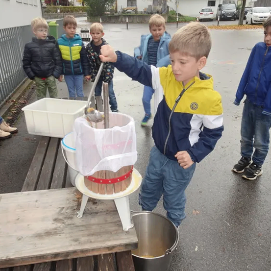 Eine Gruppe junger Jungen ist im Freien. Ein Junge benutzt ein Werkzeug, um eine Holzpresse zu drücken, unter der sich ein Metalleimer befindet.