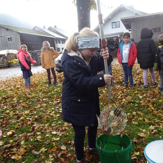 Ein Mädchen in einem blauen Mantel pflückt Äpfel mit einem langen Stock und Korb, umgeben von Kindern und gefallenen Blättern.
