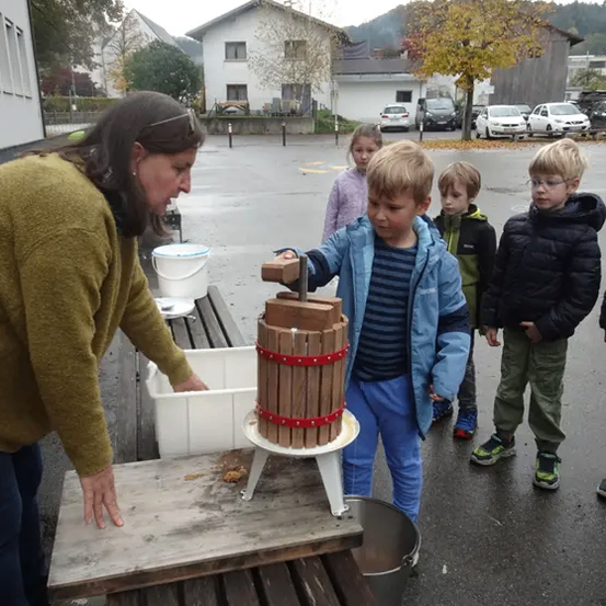 Eine Frau hilft einem Kind, einen Apfelpressen draußen zu drücken. Mehrere Kinder beobachten. Im Hintergrund sind Häuser und Autos zu sehen.