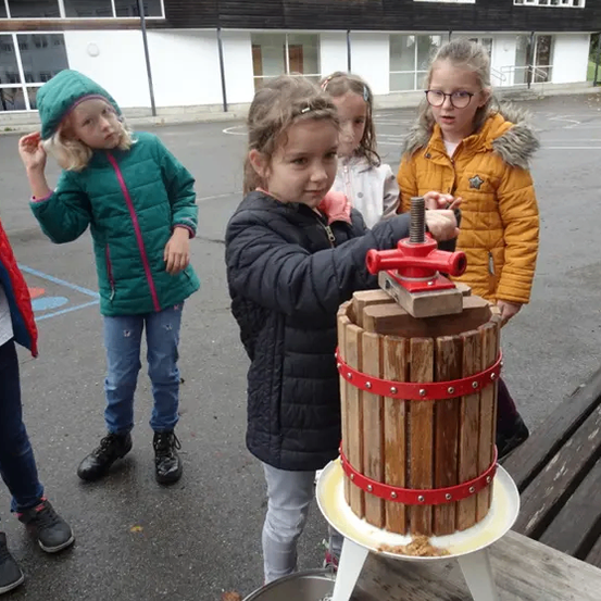Ein Mädchen bedient draußen eine hölzerne Presse, umgeben von anderen Kindern. Das Gebäude im Hintergrund hat viele Fenster.