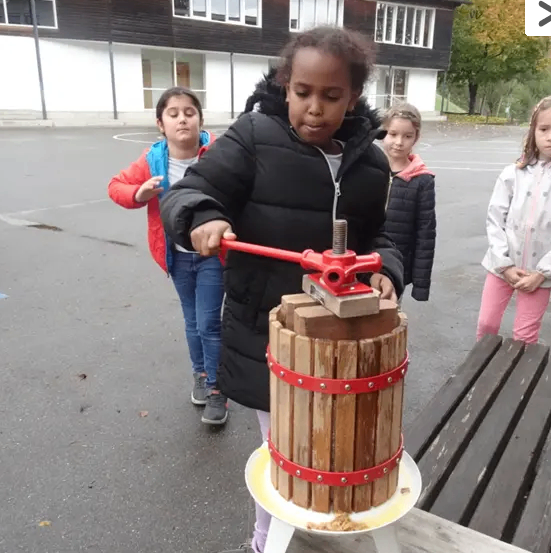 Ein junges Mädchen bedient eine Holzpresse im Freien, umgeben von drei anderen Kindern und einem Gebäude im Hintergrund.