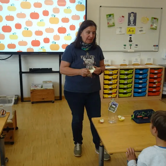 Eine Frau steht in einem Klassenzimmer und hält eine Frucht. Ein Junge sitzt an einem Tisch und schaut sie an.