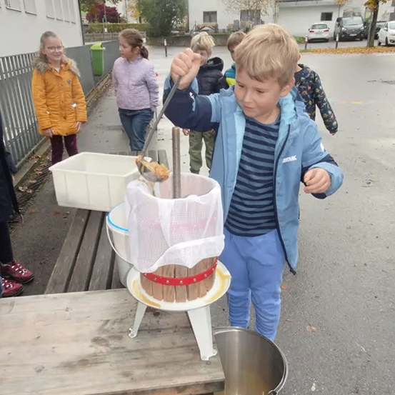 Ein Junge in einer blauen Jacke presst Trauben in einen Metalleimer. Hinter ihm beobachten Kinder. In der Nähe steht ein Tisch mit einer Weinpresse.