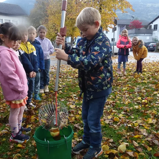 Ein junger Junge benutzt einen langen Stock, um einen Apfel aus einem Eimer voller Blätter zu pflücken. Eine Gruppe von Kindern beobachtet ihn im Garten.