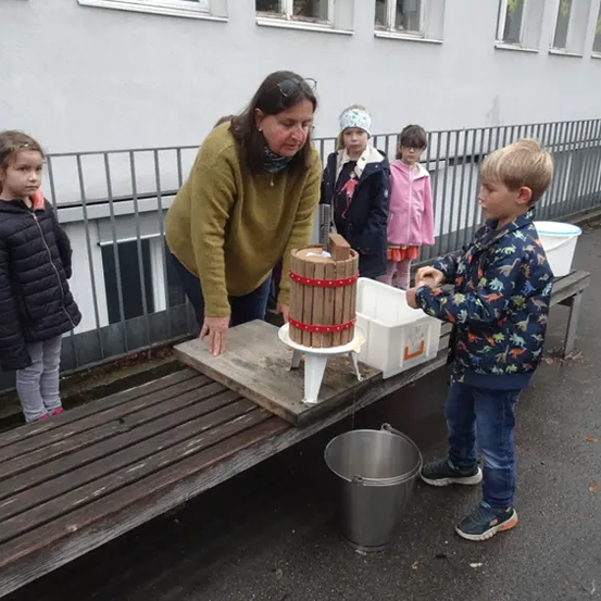 Eine Frau hilft einem Jungen mit einem Eimer bei der Verwendung einer Apfelpresse im Freien. Hinter ihnen beobachten mehrere Kinder. Ein Gebäude mit Fenstern und einem Zaun ist im Hintergrund zu sehen.