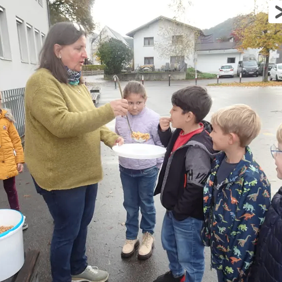 Eine Frau serviert einer Gruppe von Kindern Essen in einer Außenumgebung. Die Kinder stehen um sie herum und essen eifrig von Tellern. Die Umgebung scheint ein Schulhof zu sein, mit einem Gebäude im Hintergrund.