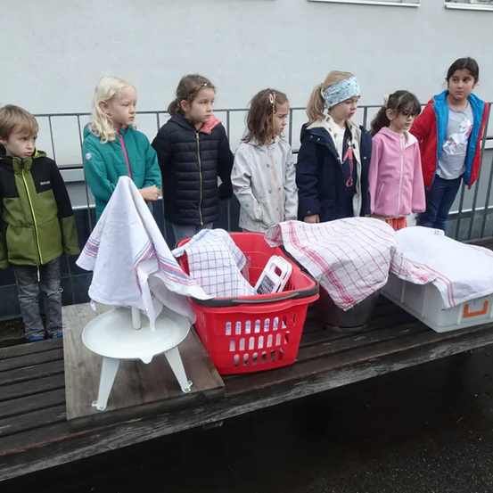 Mehrere Kinder stehen hinter einer Bank mit verschiedenen Wäschestücken, darunter ein roter Korb und weiße Aufbewahrungsboxen. Sie sind in Winterkleidung gekleidet.