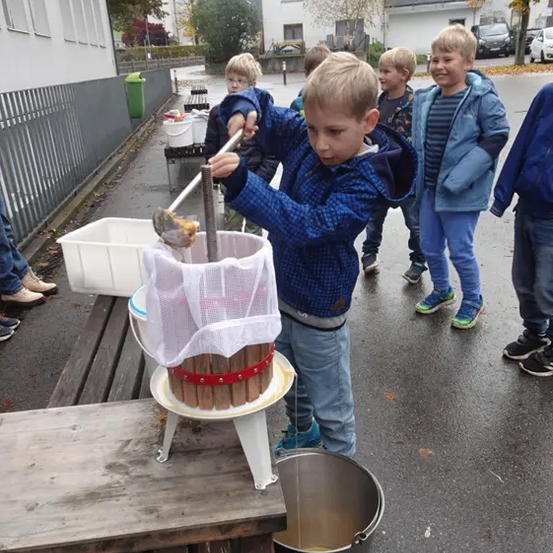 Ein junger Junge in einem blauen Mantel benutzt eine manuelle Apfelpresse. Er ist von anderen Kindern in Jacken umgeben, möglicherweise in einer Schulumgebung. Hinter ihm stehen mehrere weiße Eimer auf einem Holztisch.