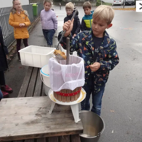 Ein junger Junge benutzt eine Presse, um Saft aus einer Frucht zu pressen, während einige Kinder zuschauen.