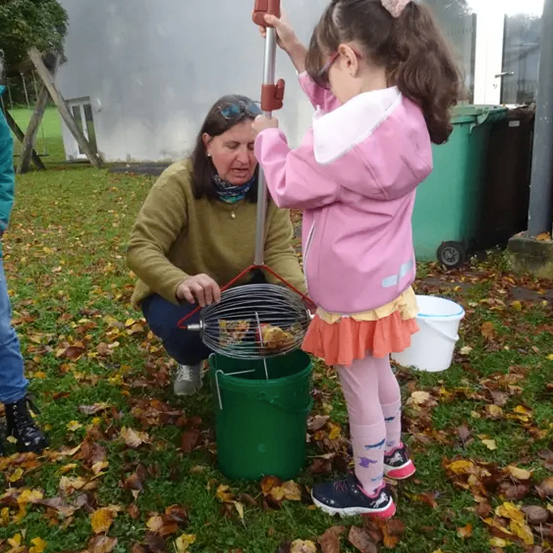 Eine Frau und ein Mädchen sind auf einem Hof, das Mädchen hält einen Metalleimer mit Essen, die Frau schaut das Mädchen an.