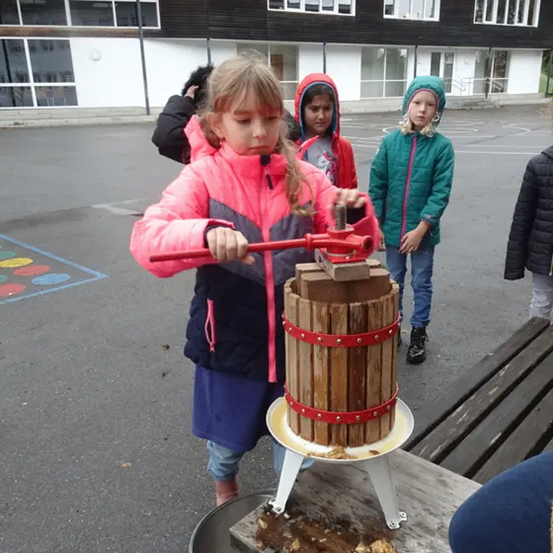 Ein kleines Mädchen bedient eine hölzerne Presse im Freien, während drei andere Kinder zuschauen. Hinter ihr steht ein Gebäude mit vielen Fenstern.
