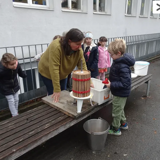 Eine Frau hilft einem jungen Jungen, eine manuelle Apfelpresse zu benutzen, während Kinder beobachten. Sie sind draußen in der Nähe eines Gebäudes.