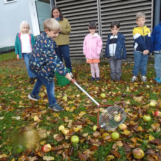 Ein junger Junge benutzt einen Obstpflücker, um Äpfel vom Boden aufzusammeln, umgeben von gefallenen Blättern. Hinter ihm stehen mehrere Kinder und eine erwachsene Frau und beobachten. Im Hintergrund befindet sich ein Gebäude mit einer Glastür.