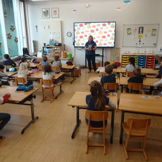 Eine Lehrerin steht vor einem Bildschirm in einem Klassenzimmer, während Schüler an ihren Tischen sitzen. Die Lehrerin scheint eine Unterrichtsstunde zu geben.