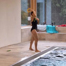 Eine Frau in einem schwarzen Badeanzug übt das Werfen eines gelben Frisbees am Pool, umgeben von einem gefliesten Boden und einem großen Glasfenster.