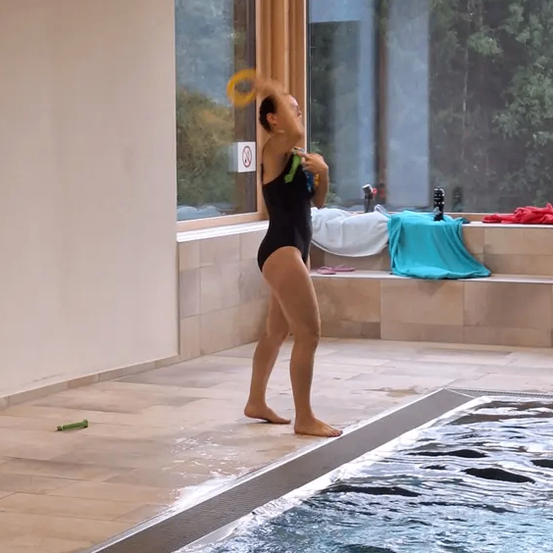 Eine Frau in einem schwarzen Badeanzug übt das Werfen eines gelben Frisbees am Pool, umgeben von einem gefliesten Boden und einem großen Glasfenster.