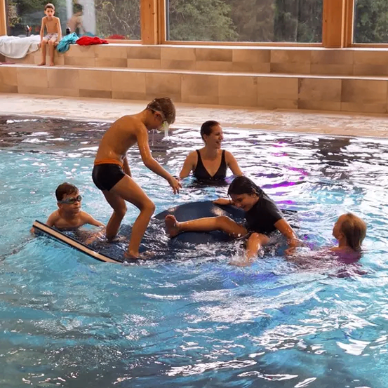 Mehrere Kinder und Erwachsene schwimmen in einem Pool. Ein junger Junge steht auf einem schwimmenden Brett, während andere darauf sitzen. Der Pool hat Glasfenster an der Seite.
