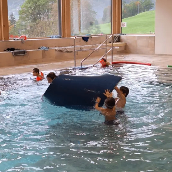 Eine Gruppe von Kindern schwimmt im Pool. Ein Kind hält sich an einem blauen Wasserrutsche fest.