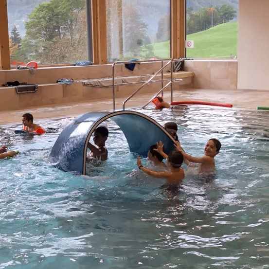 Kinder schwimmen und spielen in einem Pool mit einem blauen Bogen, während andere auf Bänken entspannen und die Aussicht genießen.