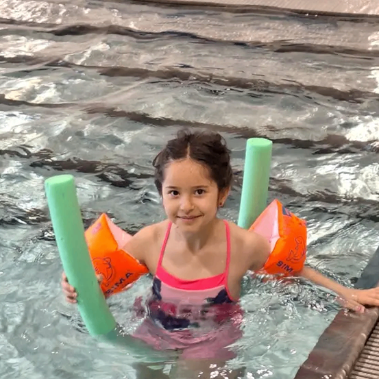 Ein kleines Mädchen mit einem rosa Badeanzug und orangefarbenen Schwimmhilfen lächelt und hält zwei grüne Rohre in der Hand, während es in einem Schwimmbecken steht.