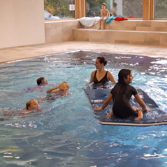 Ein Schwimmbecken mit vier Schwimmenden, zwei Erwachsenen und zwei Kindern. Eine Frau sitzt auf einer Matte, während die anderen schwimmen. Ein Junge sitzt auf der Kante. Bäume und ein Auto sind außerhalb des Beckens sichtbar.