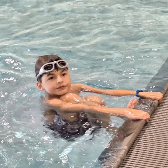 Ein junger Junge mit Schwimmbrille schwimmt im Pool. Er lehnt sich mit den Händen auf der Kante an die Seite.