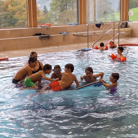 Eine Gruppe von Kindern schwimmt in einem Pool und trägt Schwimmwesten. Ein Kind hält ein Surfbrett. Der Pool hat Glaswände und bietet einen malerischen Ausblick.