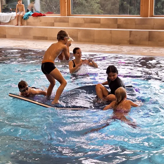 Eine Gruppe von Kindern schwimmt in einem Pool, in der Nähe von einigen Erwachsenen. Die Erwachsenen stehen am Rande des Pools. Ein Kind liegt auf einem Surfbrett.