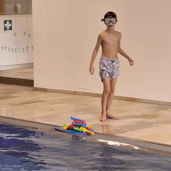 Ein junger Junge steht am Rand eines Schwimmbeckens mit Schwimmbrille und Spielzeug auf einem Teppich. Er scheint für ein Foto zu posieren.
