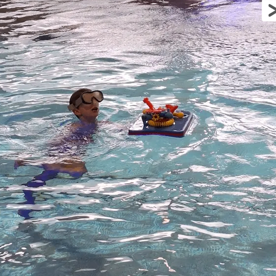 Ein junger Junge mit Schwimmbrille schwimmt in einem Pool, in dem ein Spielzeugboot schwimmt.