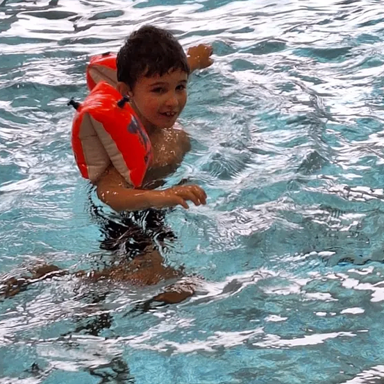 Ein junger Junge schwimmt in einem Pool mit einem orangefarbenen Rettungsweste. Seine Arme sind ausgestreckt, als ob er etwas erreichen würde.