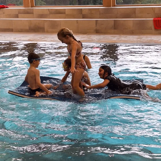 Eine Frau hilft einem jungen Mädchen und einem Jungen auf einem Surfbrett, im Pool zu schwimmen. Bäume sind durch die großen Fenster sichtbar.