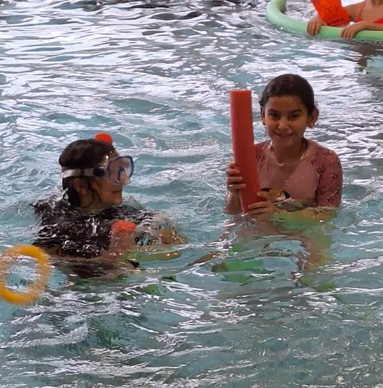 Kinder schwimmen in einem Pool, tragen Schwimmbrillen und halten Schwimmhilfen. Ein Kind trägt einen Schnorchel.
