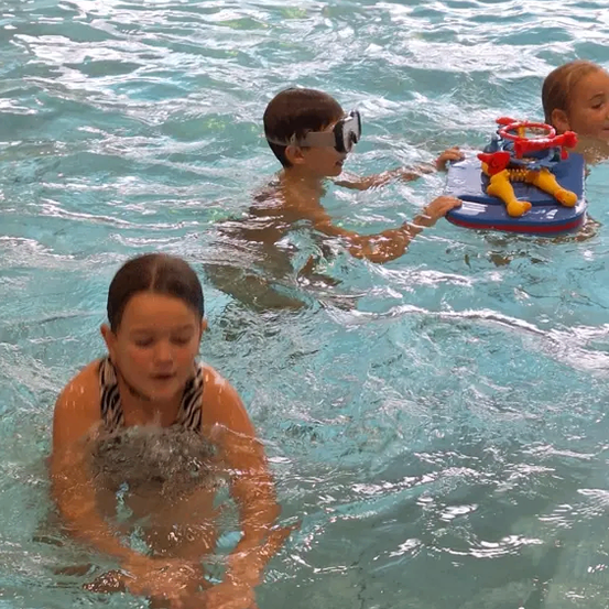 Drei Kinder schwimmen in einem Pool. Ein Mädchen in der Mitte trägt Schwimmbrillen und hält ein Spielzeug. Ein weiteres Mädchen schwimmt auf einem blauen Brett. Das dritte Kind hat langes Haar.