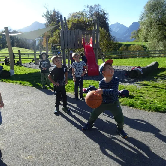 Eine Gruppe von Kindern, die an einem sonnigen Tag Basketball spielen, mit Bergen im Hintergrund.
