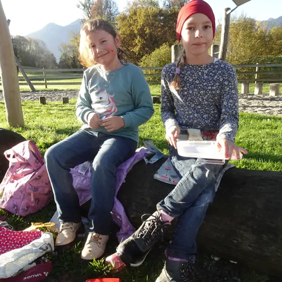 Zwei Mädchen sitzen auf einem Baumstamm in einem Park und genießen ihr Essen. Eine von ihnen trägt eine rote Mütze. Sie haben einen Rucksack, eine rote Brotdose und andere Gegenstände auf dem Boden. Im Hintergrund sieht man ein Grasfeld mit Bäumen und einem Zaun.